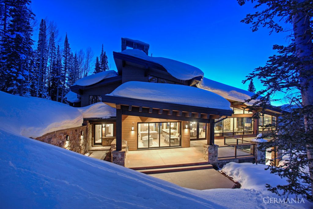 Luxury, custom-built home in Park City, UT. Covered in snow with light shining through big, beautiful windows showcasing a high end interior.