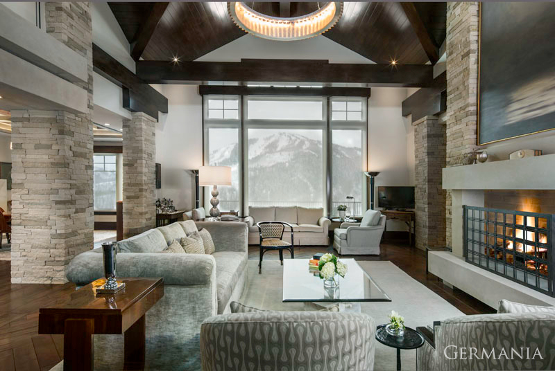 A Park City, Utah luxury custom-built home's living room. High-end grey seating, stone covered pillars, and dark wood accents fill the room.